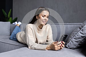 Happy young woman relax on comfortable couch at home texting messaging on smartphone, smiling girl use cellphone, browse wireless