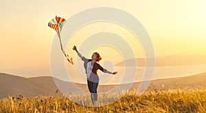 Happy young woman running with kite on glade at sunset in summer