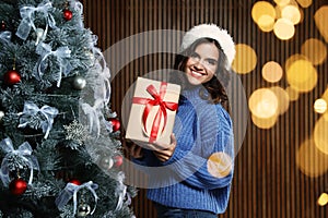 Happy young woman in Santa hat with gift near Christmas tree at home, bokeh effect