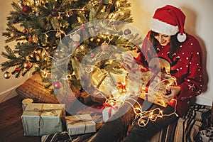 happy young woman in santa hat opening gift box at golden beautiful christmas tree with lights and presents in festive room.