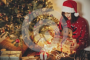 happy young woman in santa hat opening gift box at golden beautiful christmas tree with lights and presents in festive room.