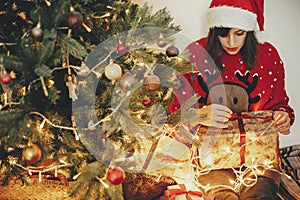 happy young woman in santa hat opening gift box at golden beautiful christmas tree with lights and presents in festive room.