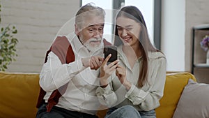 Happy young woman and senior grandfather web surfing in social media on smartphone, watching news online and laughing