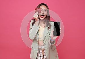 Happy young woman showing credit card and mobile phone standing over pink background, Online payment, hands holding credit card
