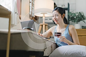 A happy young woman is shown shopping online using her laptop and credit card at home.