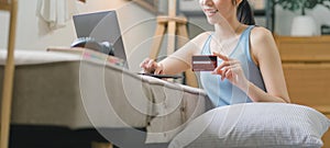 A happy young woman is shown shopping online using her laptop and credit card at home.