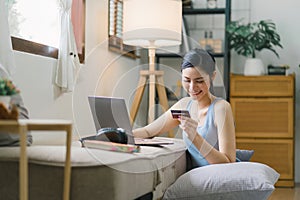 A happy young woman is shown shopping online using her laptop and credit card at home.