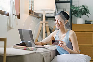 A happy young woman is shown shopping online using her laptop and credit card at home.