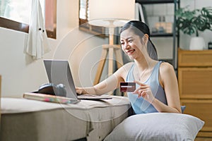 A happy young woman is shown shopping online using her laptop and credit card at home.