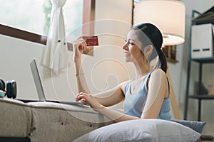 A happy young woman is shown shopping online using her laptop and credit card at home.