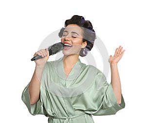 Happy young woman in silk bathrobe with hair curlers singing into hairbrush on white background