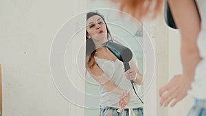 Happy young woman singing in bedroom, looking at mirror at reflection. Girl uses hair dryer and dances. Slow motion.