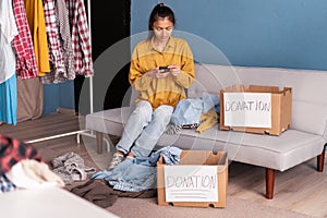 Happy young woman sit on couch stuck clothes in donation box at home, caring female volunteer put apparel in carton