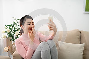 Happy young woman sit on sofa, holding phone looking at screen waving hand video calling distance friend