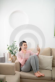 Happy young woman sit on sofa, holding phone looking at screen waving hand video calling distance friend