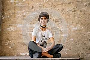 A happy young woman sits in a white t- shirt with the words we should all be feminists, concept