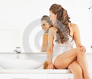 Happy young woman sitting in bathroom and looking in mirror