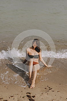 Happy young woman sitting on beach with waves. Stylish tanned  girl in modern swimsuit relaxing on seashore. Summer vacation.