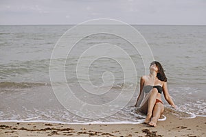 Happy young woman sitting on beach with waves. Stylish tanned  girl in modern swimsuit relaxing on seashore. Summer vacation.