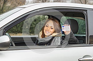 Happy young woman sitting in the car smiling at the camera showing her driver license