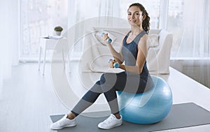 Lovely young woman sitting on fitness ball and exercising with dumbbells at home, empty space