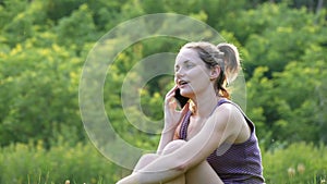 Happy Young Woman Sitting on Green Lawn and Talking on the Phone or Smartphone