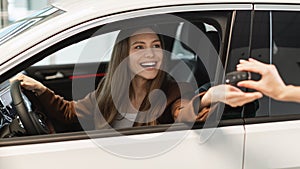 Happy young woman sitting inside modern auto, taking car key from salesperson, purchasing new vehicle at dealership