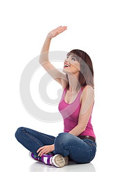 Happy Young Woman Is Sitting Legs Crossed On Floor, Looking Up And Waving Hand