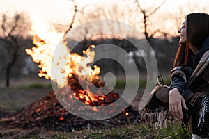 Happy young woman sitting near the evening midspring night campfire among field