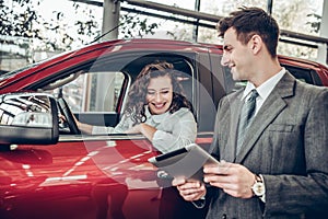 Happy young woman sitting in a new car at car salon professional salesman showing them information on digital tablet couple buying