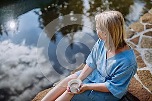 Happy young woman sitting by pond near cottege and enjoying cup of morning coffee on summer vacation in mountains.