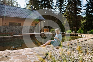 Happy young woman sitting by pond near cottege and enjoying cup of morning coffee on summer vacation in mountains. High