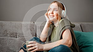Happy young woman sitting on sofa with in headphones with cup of tea. Woman or teen girl resting, bliss enjoy listening to music