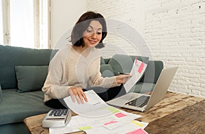 Happy young woman sitting on sofa surrounded by papers calculating expenses and paying bills