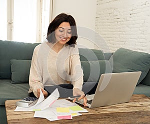 Happy young woman sitting on sofa surrounded by papers calculating expenses and paying bills