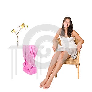 Happy young woman sitting on a wicker chair beside a table with a beautiful flower in a vase and a pink towel