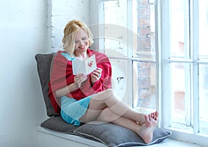Happy young woman sitting on windowsill with love postcard