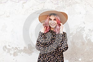 Happy young woman with smile with glamour pink hair in fashion vintage straw hat in black stylish beautiful floral dress near wall