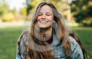 Happy young woman smiling broadly with healthy toothy smile, wearing denim jacket, posing on nature background in the park. People