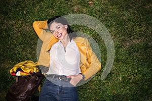 Happy young woman smiling and lying on green grass wearing colorful outfit with yellow jacket and blue jeans at night