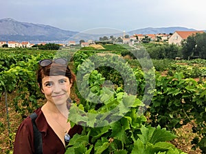 A happy young woman standing amongst the sprawling wine vineyard growing the local grk grapes with the small town of Lumbarda