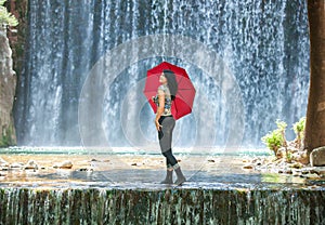 Happy young woman standing in a creek with a red umbrella in front of an impressive water fall