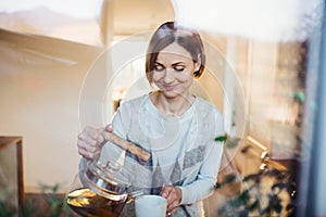 A young woman standing indoors in kitchen, pouring tea. Shot through glass.