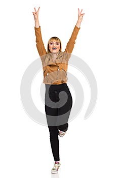 Happy Young Woman Is Standing On One Leg With Arms Raised And Showing Peace Hand Sign