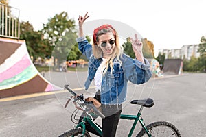 Happy young woman stands with a bicycle on the background of a skate park and rejoices with raised hands. Joyful hipster girl
