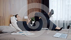 Happy young woman in suit listening to music wearing headphones in hotel bed