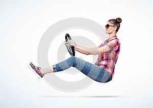 Happy young woman in sunglasses and colored plaid shirt rides a car holding a steering wheel in her hands, on light background