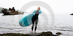 Happy young woman sup surfer in wetsuit posing with surfboard at damaged antique wooden ship