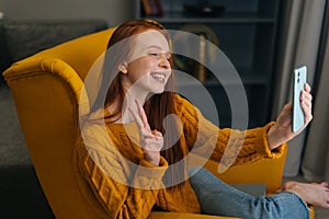 Happy young woman taking selfie picture or chatting by video call showing victory sign on mobile phone sitting in cozy
