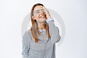Happy young woman touching and covering half of face, laughing sincere, having fun and feeling positive, standing upbeat